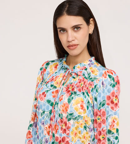 A woman with long dark hair wears the Lizzie Blue Floral Jersey Blouse, featuring a tie neck and frill stand collar, as she gazes neutrally at the camera against a plain light background.