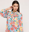 A woman with long dark hair wears the Lizzie Blue Floral Jersey Blouse, a colorful long-sleeve top with a frill stand collar and tie neck, gently touching the back of her neck while looking down against a light background.