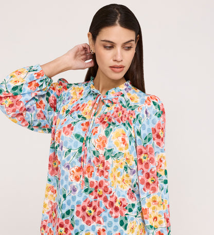 A woman with long dark hair wears the Lizzie Blue Floral Jersey Blouse, a colorful long-sleeve top with a frill stand collar and tie neck, gently touching the back of her neck while looking down against a light background.