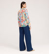 A woman with straight, dark hair wears the Lizzie Blue Floral Jersey Blouse, featuring long sleeves and a tie neck, paired with wide-leg blue jeans and beige shoes. She stands facing away against a plain light background.