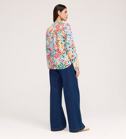 A woman with straight, dark hair wears the Lizzie Blue Floral Jersey Blouse, featuring long sleeves and a tie neck, paired with wide-leg blue jeans and beige shoes. She stands facing away against a plain light background.