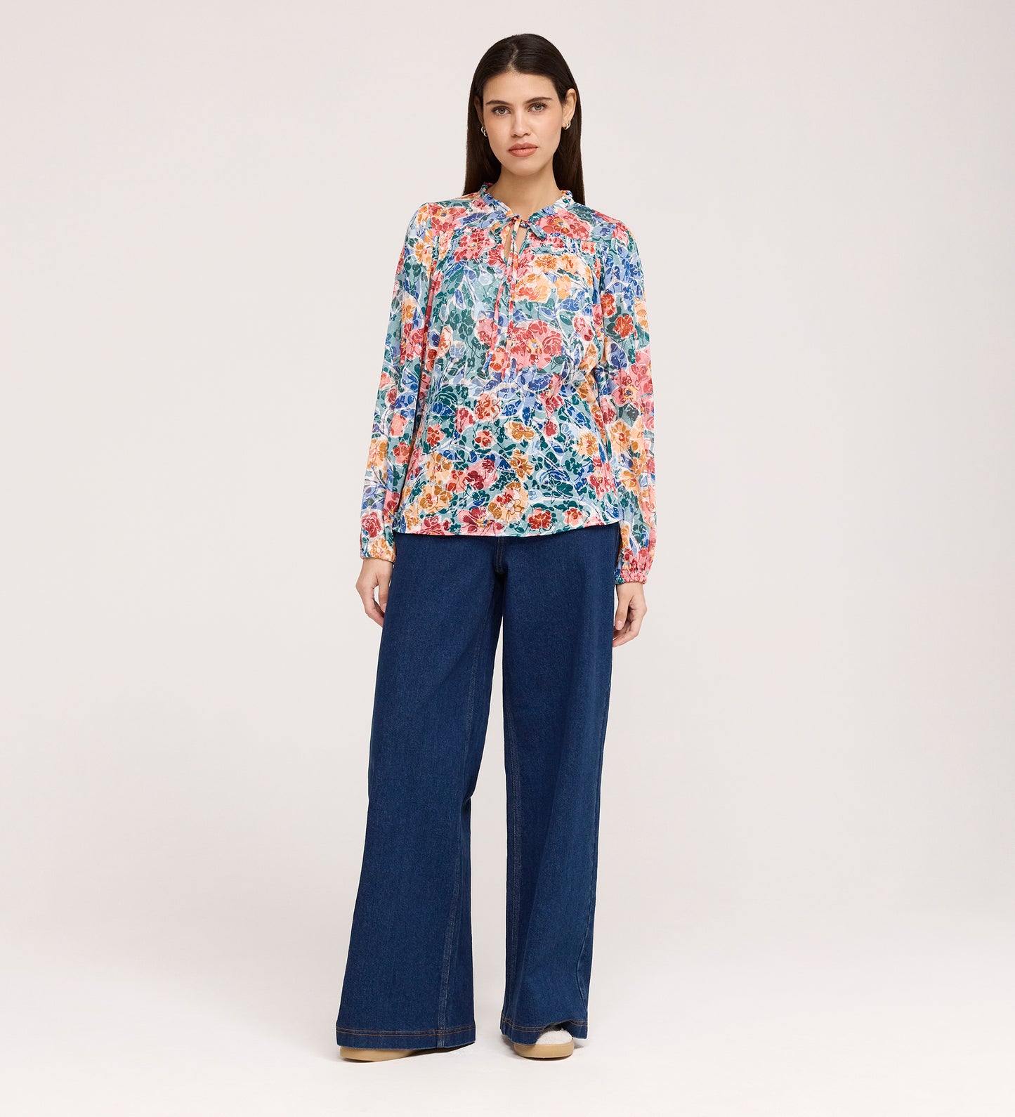 A woman stands facing forward against a plain white background, wearing the Lizzie Green Floral Jersey Blouse with long sleeves and a tie-neck, paired with wide-leg blue jeans and beige shoes.