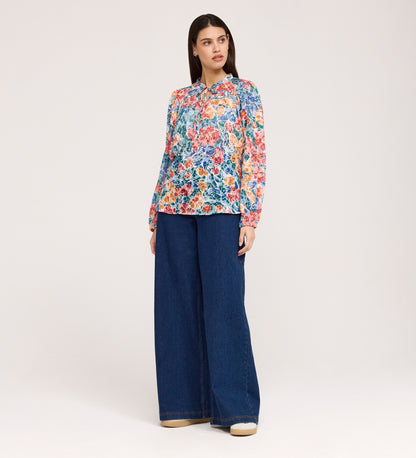 A woman with straight dark hair and a neutral expression stands against a plain background, wearing the Lizzie Green Floral Jersey Blouse paired with wide-leg blue jeans.