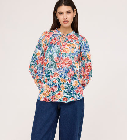 A woman wearing the Lizzie Green Floral Jersey Blouse with long sleeves stands against a plain, light background, pairing it with loose blue jeans.