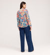 A woman faces away, wearing the Lizzie Green Floral Jersey Blouse with a stylish tie neck and wide-leg blue jeans, set against a plain light background.