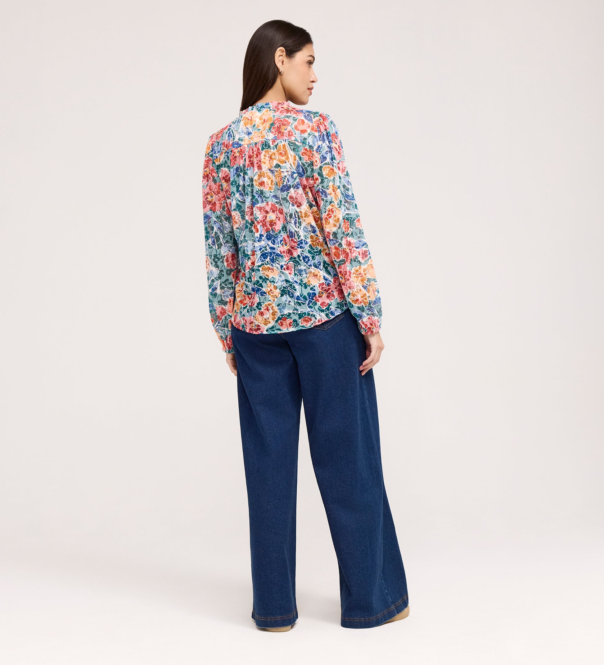 A woman faces away, wearing the Lizzie Green Floral Jersey Blouse with a stylish tie neck and wide-leg blue jeans, set against a plain light background.