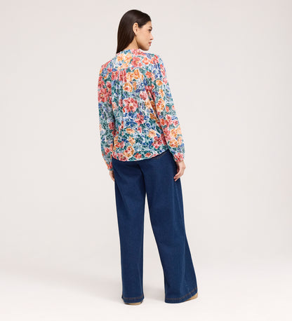 A woman faces away, wearing the Lizzie Green Floral Jersey Blouse with a stylish tie neck and wide-leg blue jeans, set against a plain light background.