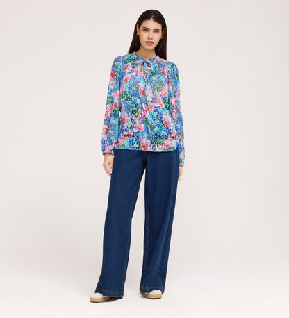 A woman with long brown hair and a neutral expression stands against a plain background, wearing the Lizzie Indigo Floral Jersey Blouse with blouson sleeves, wide-leg blue jeans, and beige shoes.
