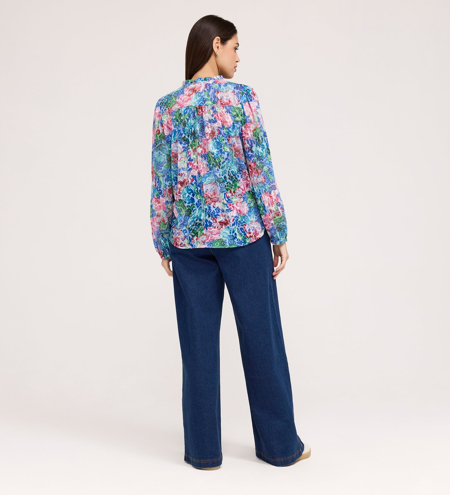 A woman stands with her back to the camera, wearing the Lizzie Indigo Floral Jersey Blouse featuring blue, pink, and purple florals, paired with wide-leg blue jeans and tan shoes against a plain light background.