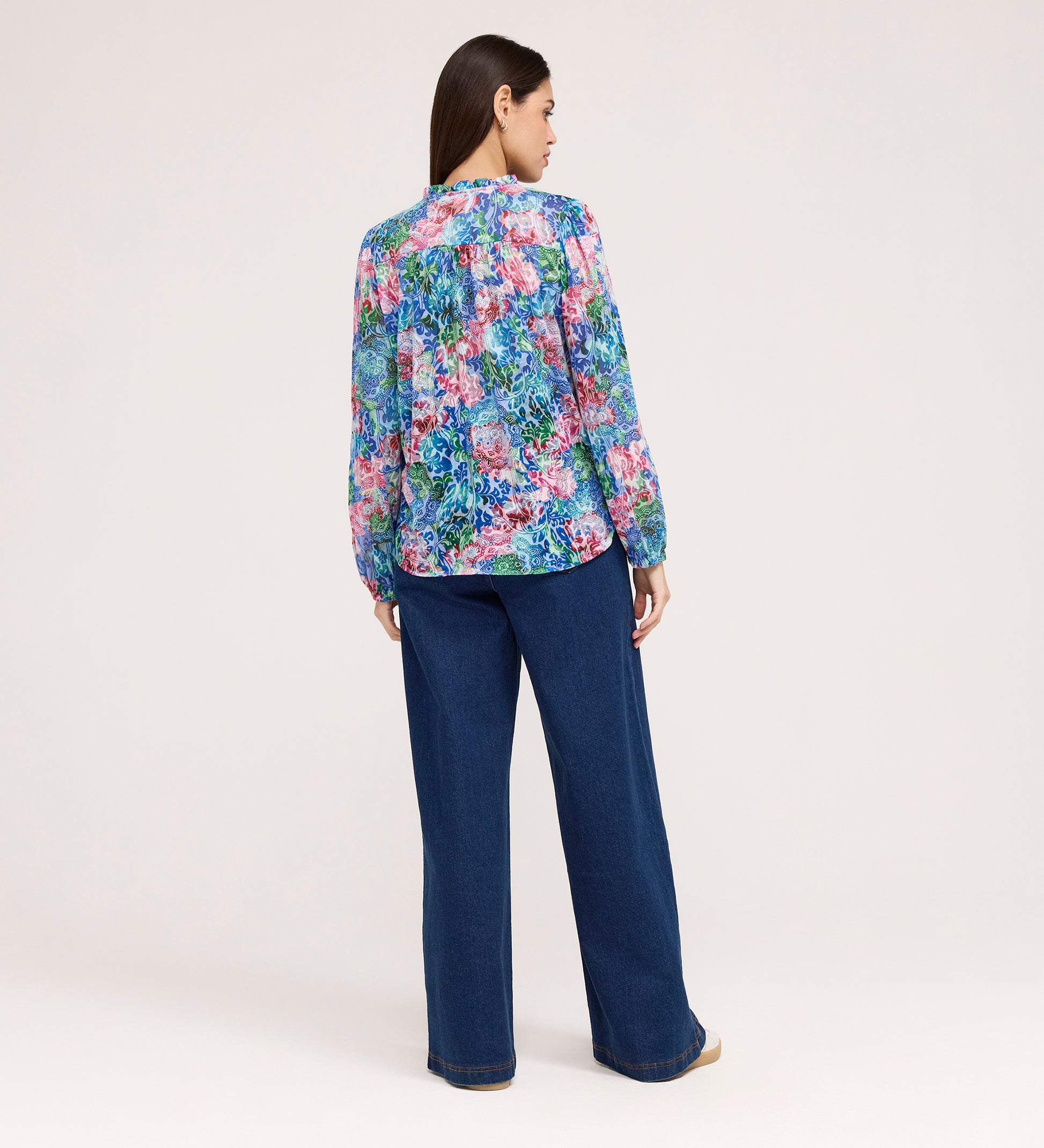 A woman stands with her back to the camera, wearing the Lizzie Indigo Floral Jersey Blouse featuring blue, pink, and purple florals, paired with wide-leg blue jeans and tan shoes against a plain light background.