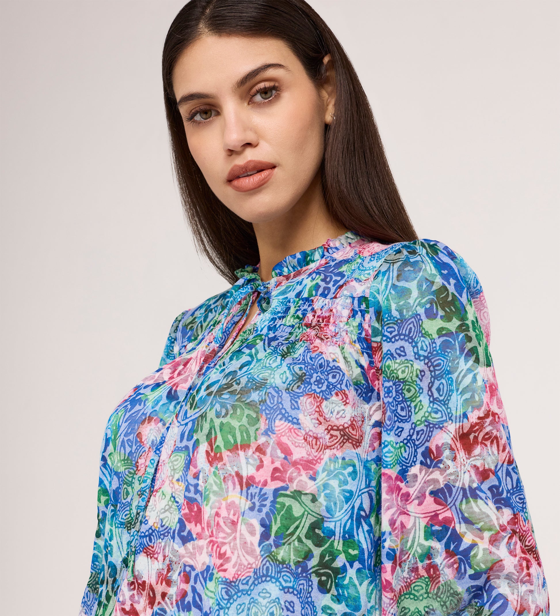 A woman with straight dark hair wears the Lizzie Indigo Floral Jersey Blouse, featuring a vibrant floral pattern and tie neck in blue, pink, green, and red. She stands against a plain light background, looking calmly at the camera.