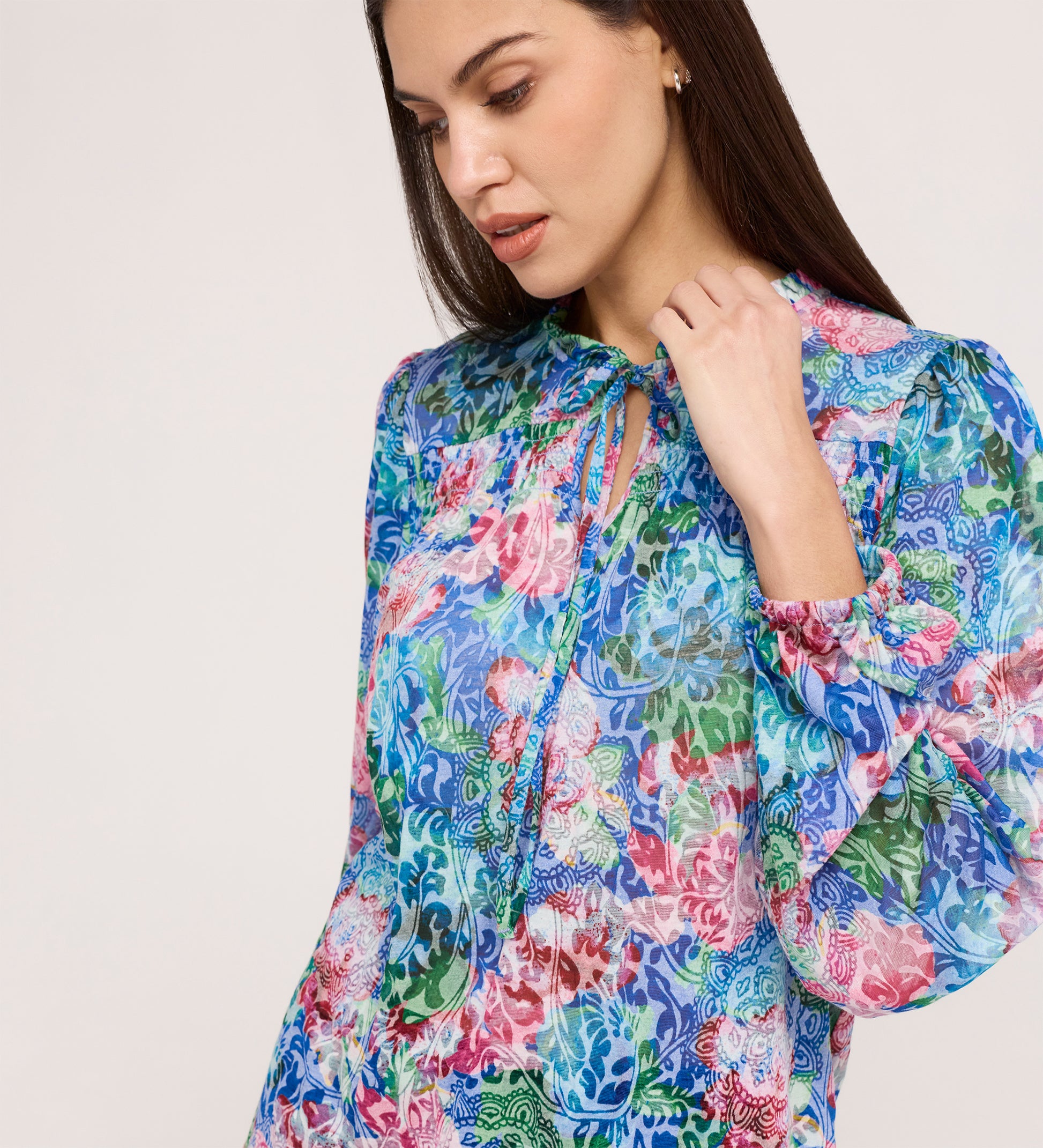 A woman with long dark hair wears the Lizzie Indigo Floral Jersey Blouse, featuring a colorful floral pattern, tie neck, and blouson sleeves. She looks down gently, touching the tie against a pale plain background.