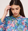 A woman with long, straight brown hair looks down, touching her head while wearing the Lizzie Indigo Floral Jersey Blouse, a tie-neck top with a colorful floral pattern, ruffled details, and blouson sleeves for a stylish touch.