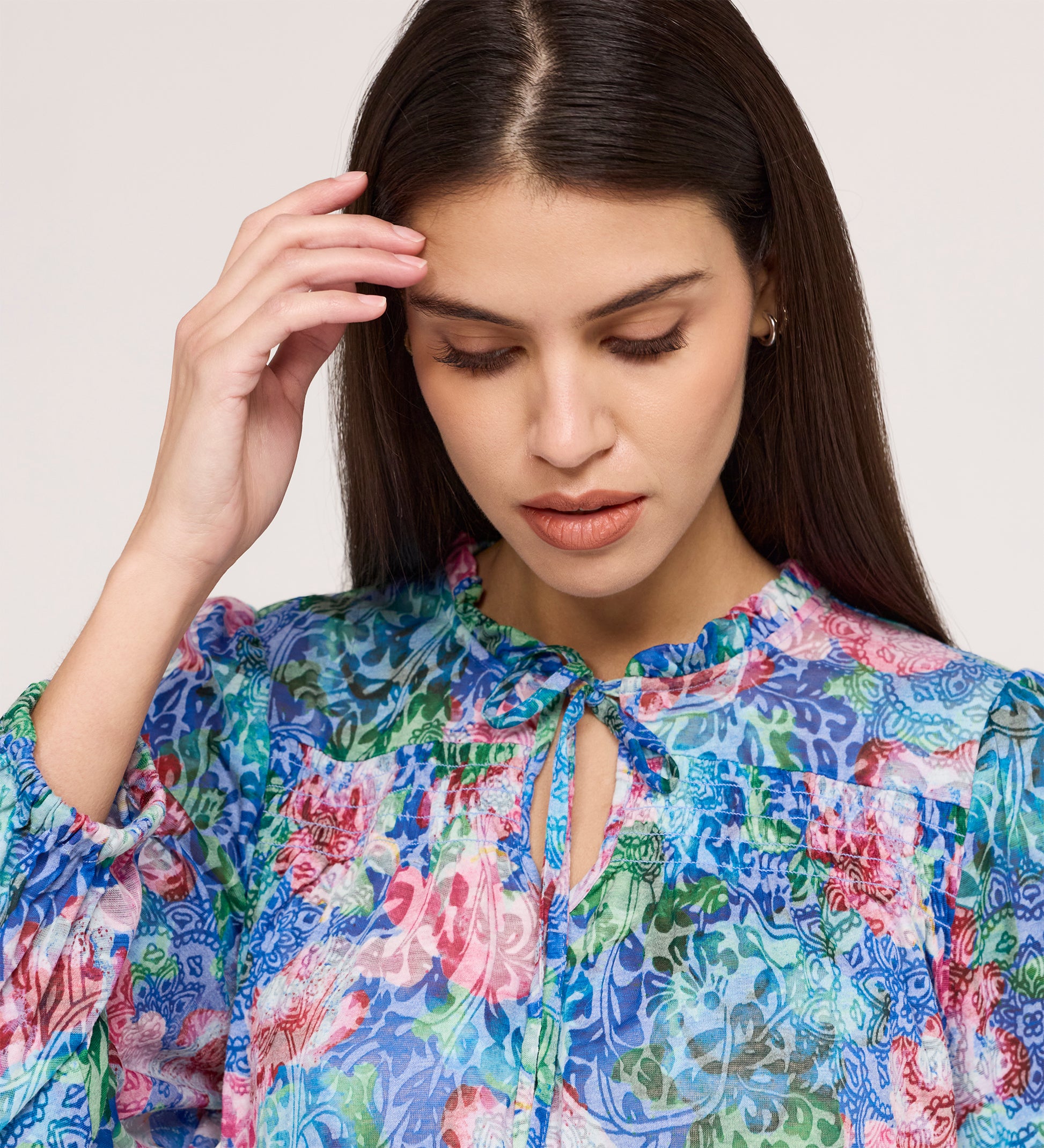 A woman with long, straight brown hair looks down, touching her head while wearing the Lizzie Indigo Floral Jersey Blouse, a tie-neck top with a colorful floral pattern, ruffled details, and blouson sleeves for a stylish touch.