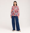 A woman stands facing forward in the Lizzie Pink Floral Jersey Blouse with blouson sleeves and wide-leg blue jeans. She has straight brown hair, neutral-toned shoes, and is pictured against a plain light background.