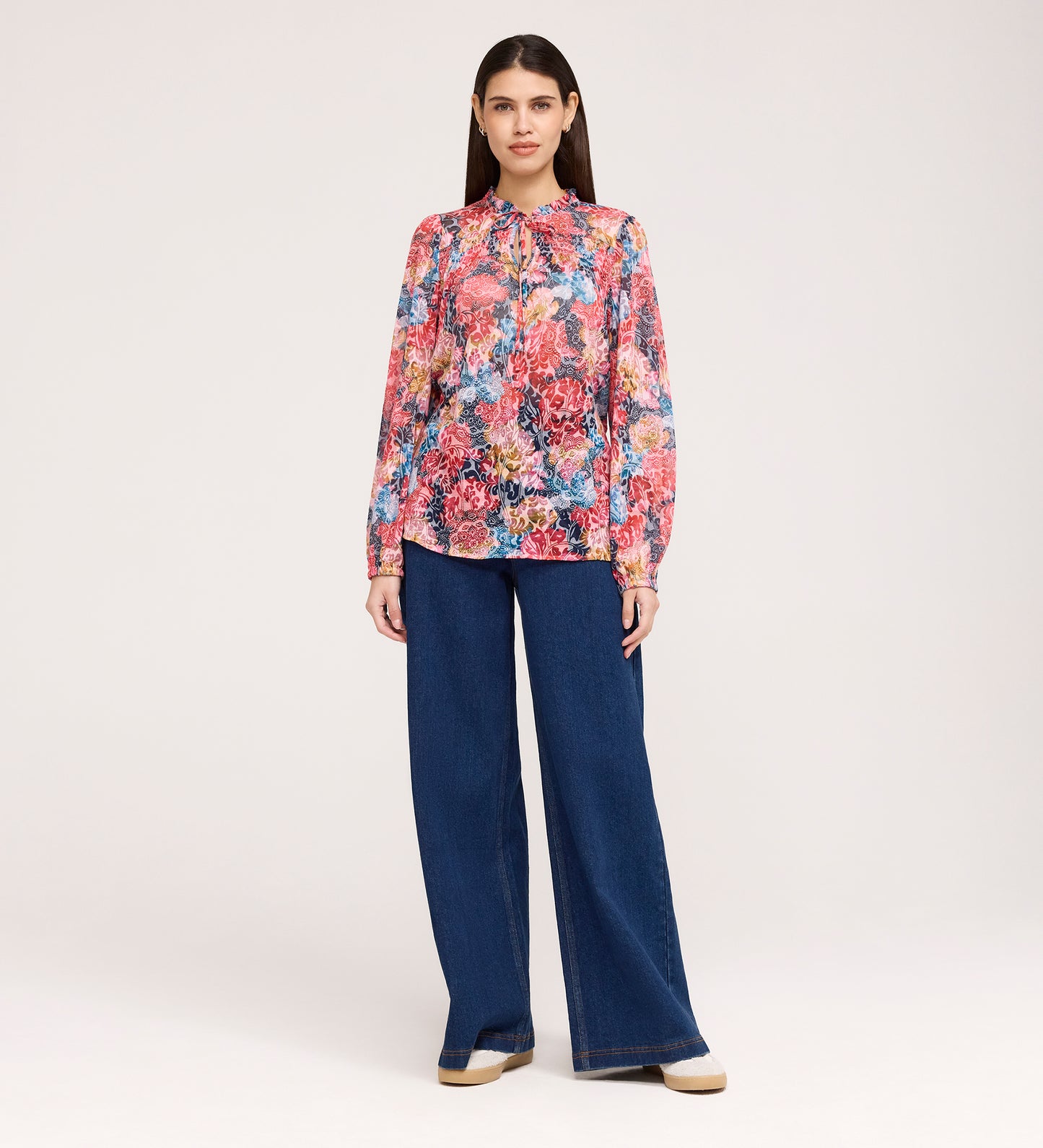 A woman stands facing forward in the Lizzie Pink Floral Jersey Blouse with blouson sleeves and wide-leg blue jeans. She has straight brown hair, neutral-toned shoes, and is pictured against a plain light background.