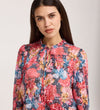 A woman with long dark hair wears the Lizzie Pink Floral Jersey Blouse, featuring a tie neck and keyhole neckline, as she stands against a plain background looking directly at the camera with a neutral expression.