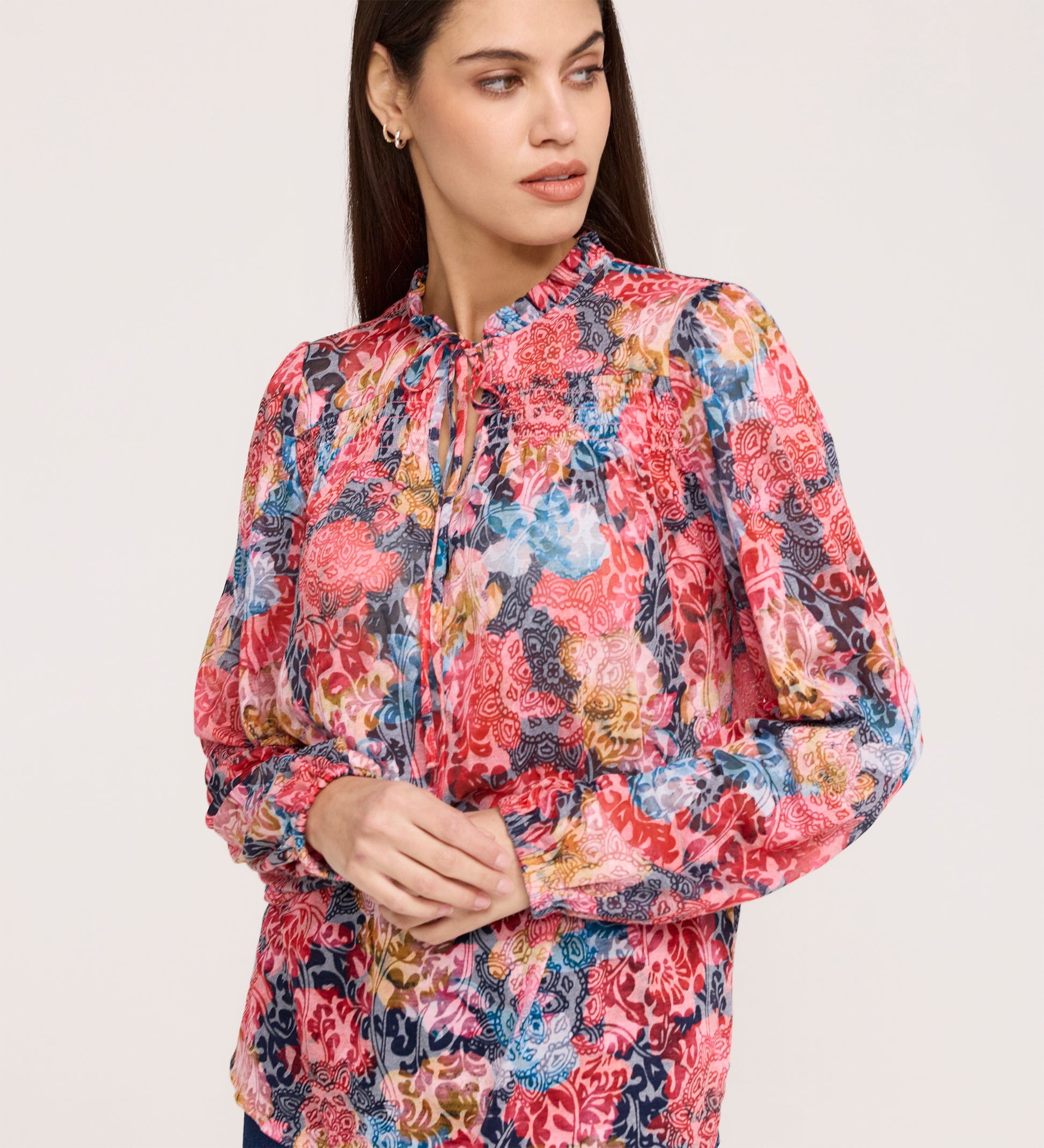A woman with long dark hair wears the Lizzie Pink Floral Jersey Blouse, featuring blouson sleeves and a tie neckline. She looks to her left with a neutral expression against a plain background.