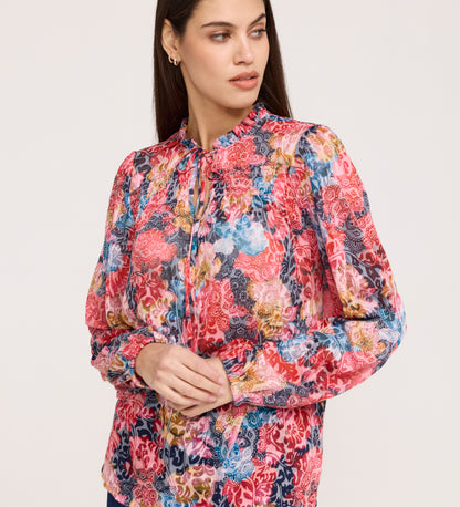 A woman with long dark hair wears the Lizzie Pink Floral Jersey Blouse, featuring blouson sleeves and a tie neckline. She looks to her left with a neutral expression against a plain background.
