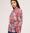 A woman with long dark hair wears the Lizzie Pink Floral Jersey Blouse, standing against a plain light background and looking slightly to the side with a subtle smile.