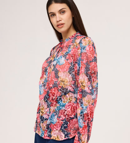 A woman with long dark hair wears the Lizzie Pink Floral Jersey Blouse, standing against a plain light background and looking slightly to the side with a subtle smile.