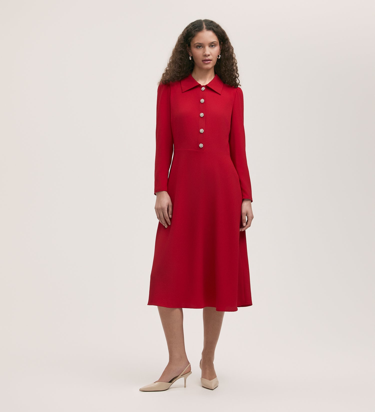 A woman with curly hair models the Lucia Red Crepe Midi Dress, featuring large buttons and a pointed collar, paired with beige low-heeled shoes against a plain light background.