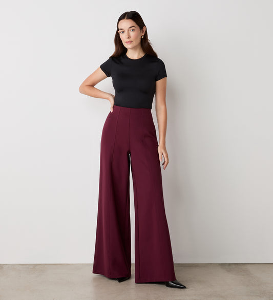A woman poses against a plain wall, wearing a fitted black t-shirt and the Matilda Burgundy Ponte Jersey Wide Leg Trousers with an elasticated waistband, paired with black pointed shoes.