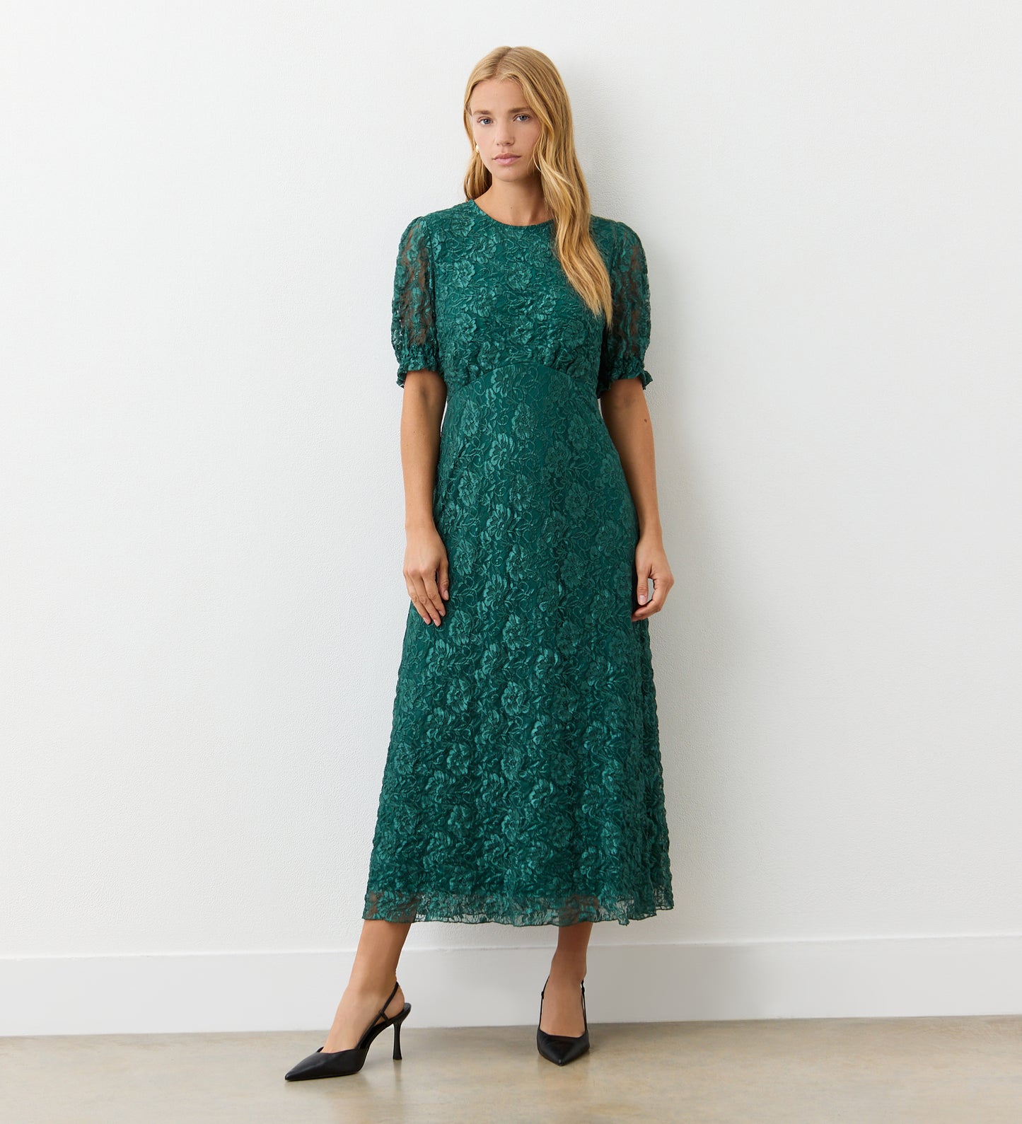 A woman stands against a white wall wearing the Mela Green Lace Midi Dress, featuring short puff sleeves and an empire line waist. She has long blonde hair and pairs the dress with black pointed-toe heels.