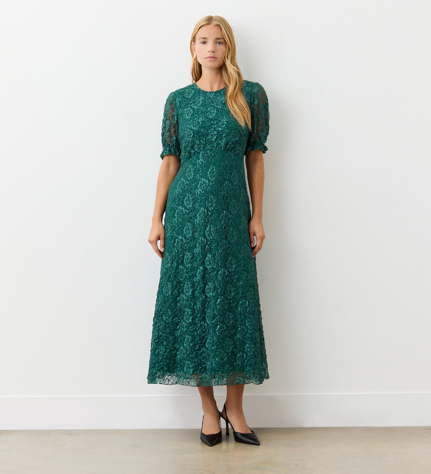 A woman stands indoors against a white wall, wearing the Mela Green Lace Midi Dress featuring an elegant neckline and short puff sleeves. She completes her look with black pointed-toe heels, her long straight hair framing her neutral expression.