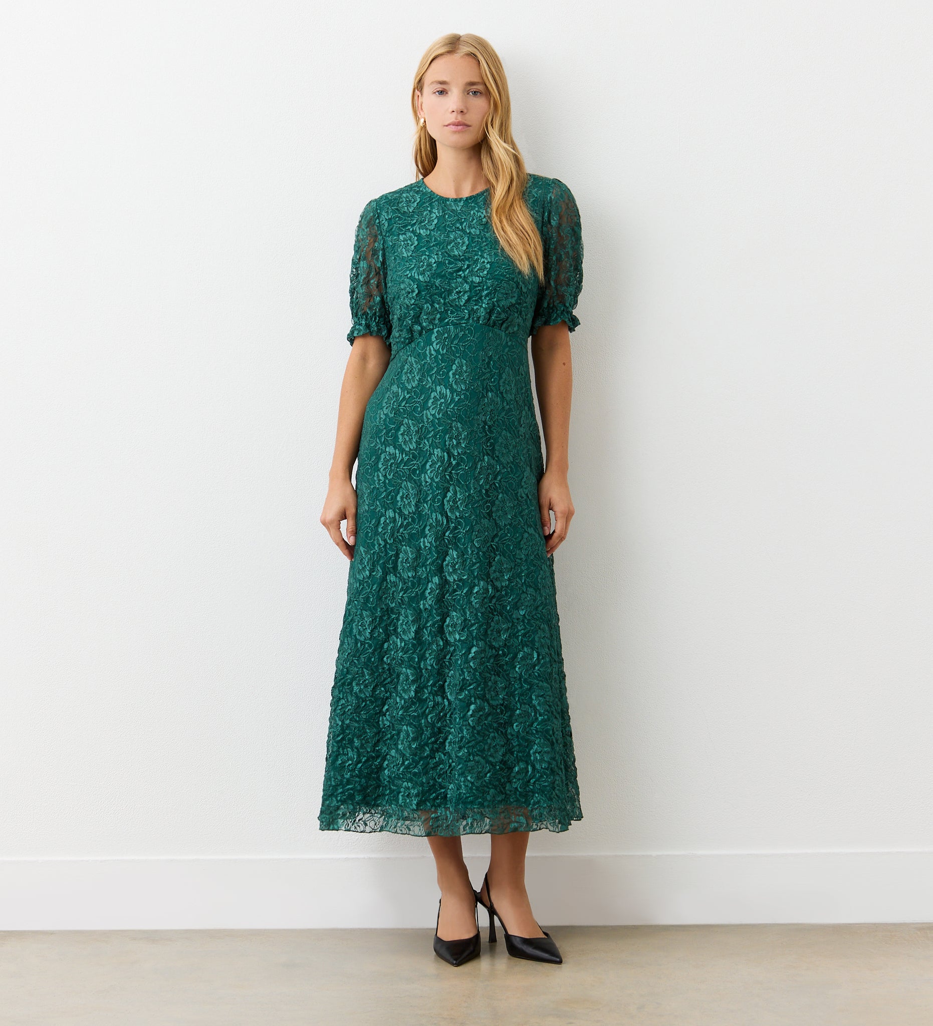 A woman stands indoors against a white wall, wearing the Mela Green Lace Midi Dress featuring an elegant neckline and short puff sleeves. She completes her look with black pointed-toe heels, her long straight hair framing her neutral expression.