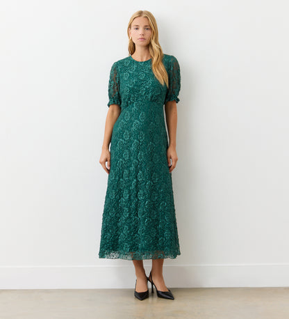 A woman stands indoors against a white wall, wearing the Mela Green Lace Midi Dress featuring an elegant neckline and short puff sleeves. She completes her look with black pointed-toe heels, her long straight hair framing her neutral expression.