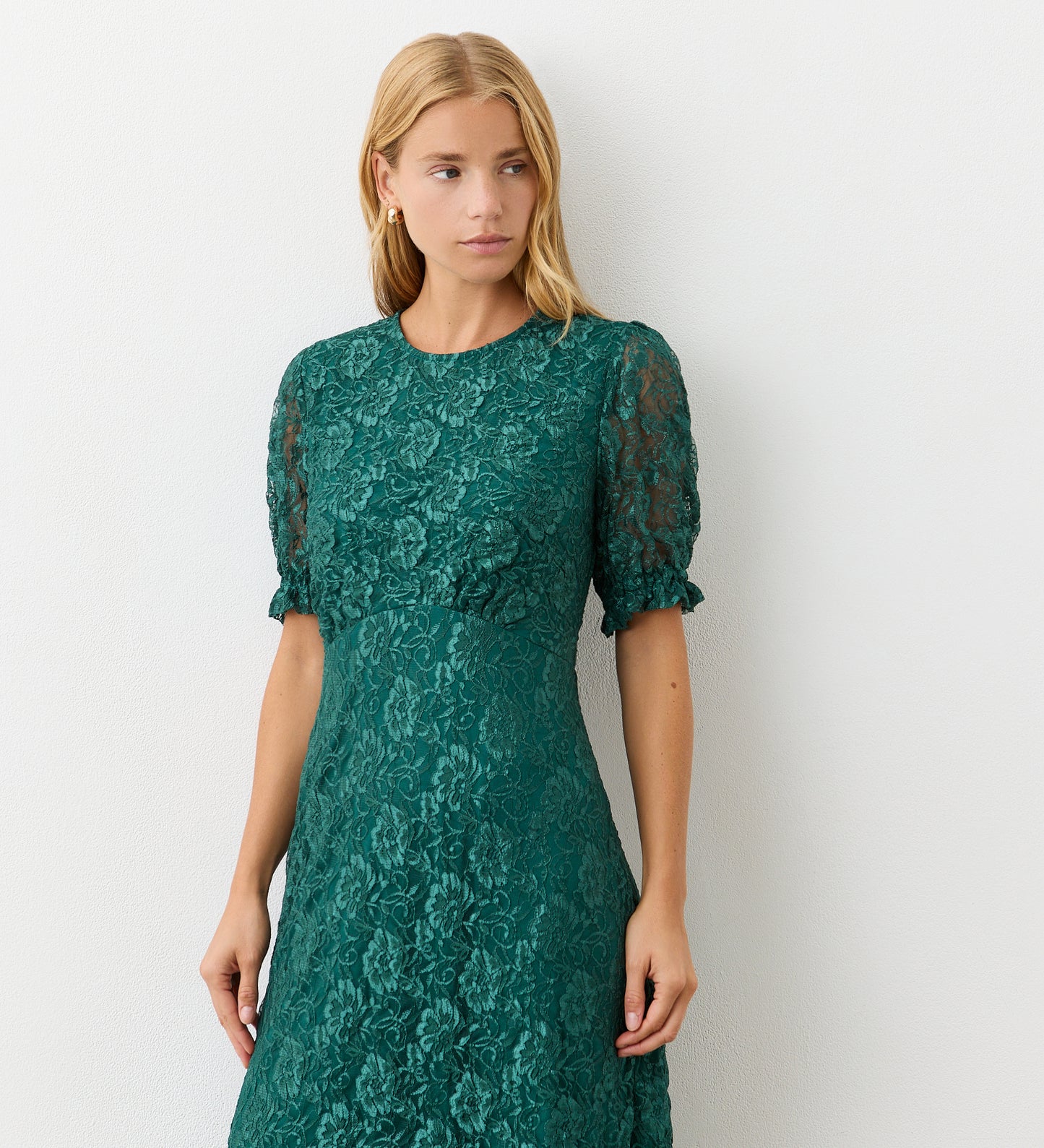 A woman with long blonde hair models the Mela Green Lace Midi Dress—featuring short sleeves and an elegant neckline—while standing against a plain white wall, looking slightly to the side with a neutral expression.