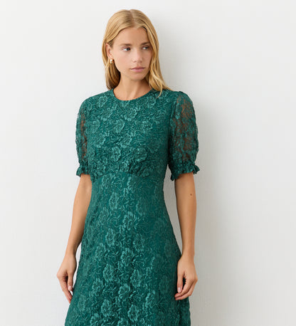 A woman with long blonde hair models the Mela Green Lace Midi Dress—featuring short sleeves and an elegant neckline—while standing against a plain white wall, looking slightly to the side with a neutral expression.