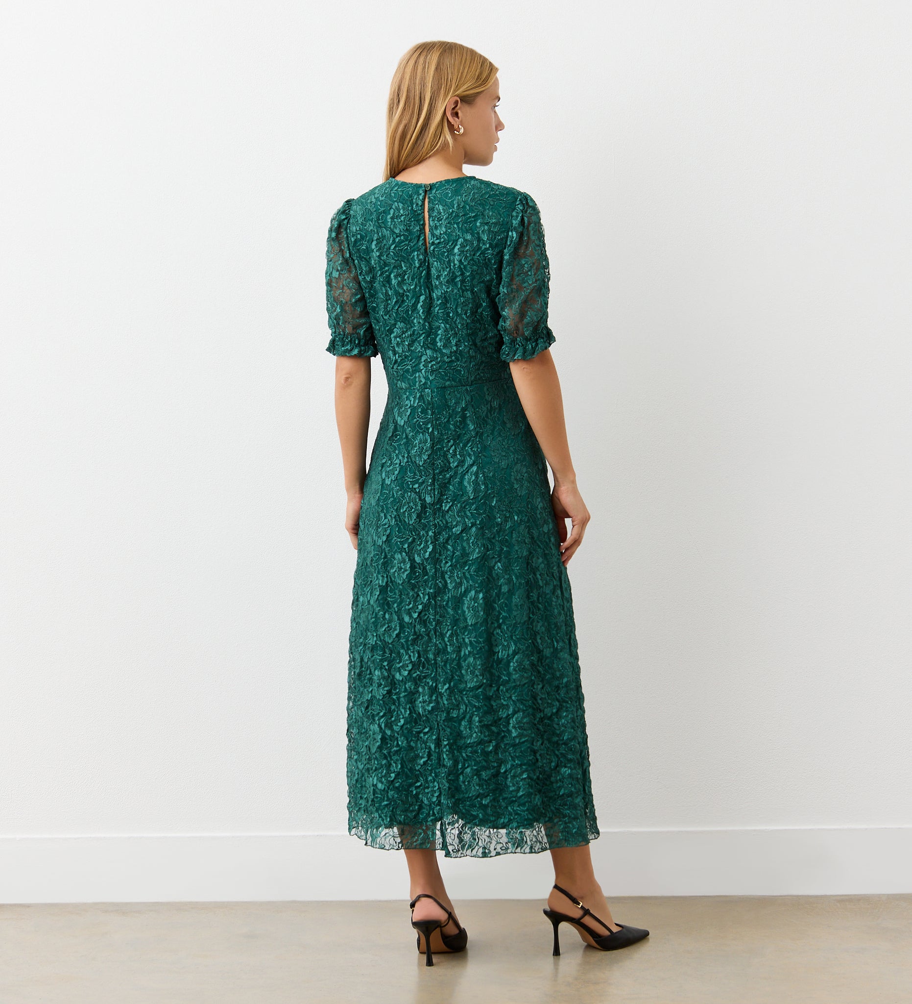 A woman with long blonde hair faces away, wearing the Mela Green Lace Midi Dress with an elegant neckline and short sleeves, paired with black heels. She stands against a plain white wall on a light-colored floor.