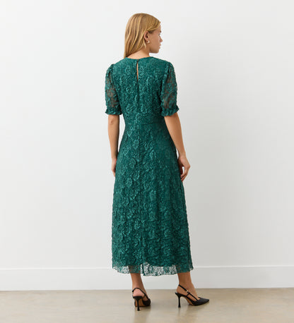 A woman with long blonde hair faces away, wearing the Mela Green Lace Midi Dress with an elegant neckline and short sleeves, paired with black heels. She stands against a plain white wall on a light-colored floor.