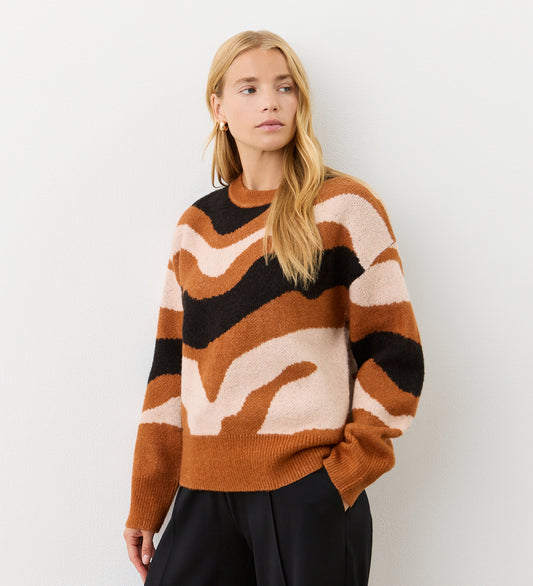 A woman with long blonde hair wears the Minnie Brown Abstract Print Knitted Jumper, featuring a brown, black, and cream pattern, paired with black pants against a plain white background.
