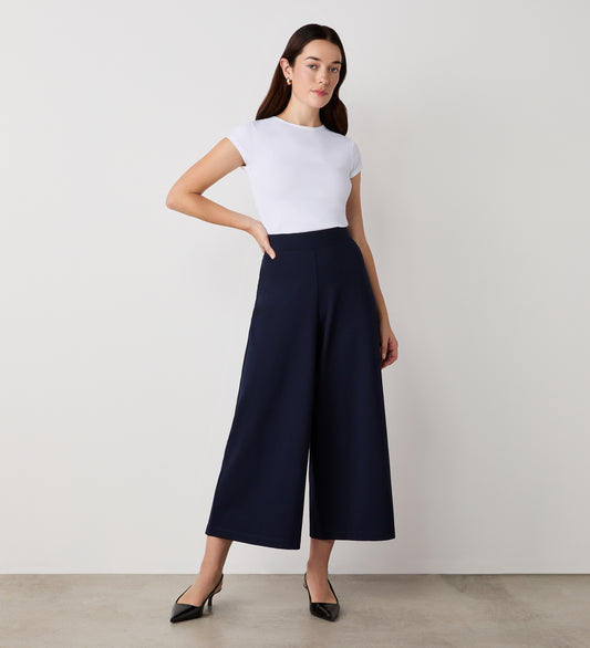 A woman with long dark hair stands against a plain wall, wearing a fitted white short-sleeve top and Miribel Navy Cropped Ponte Jersey Trousers with a high waist. She completes the look with black pointed-toe heels, one hand resting on her hip.