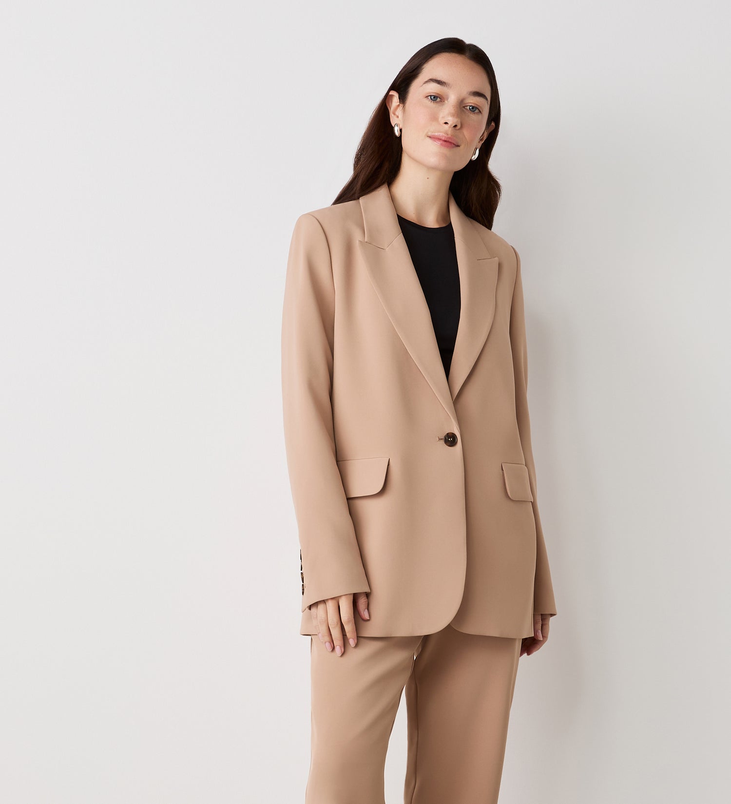 A woman with long brown hair stands against a white background, wearing the Myla Camel Longline Blazer over a black top and matching trousers. She has a neutral expression, with her hand resting by her side.
