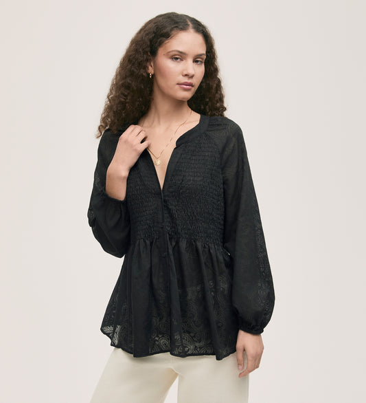 A woman with long curly hair wears the Martha Black Chiffon Blouse, featuring long sleeves, a shirred bodice, and subtle paisley burnout chiffon, paired with white pants against a plain light background.