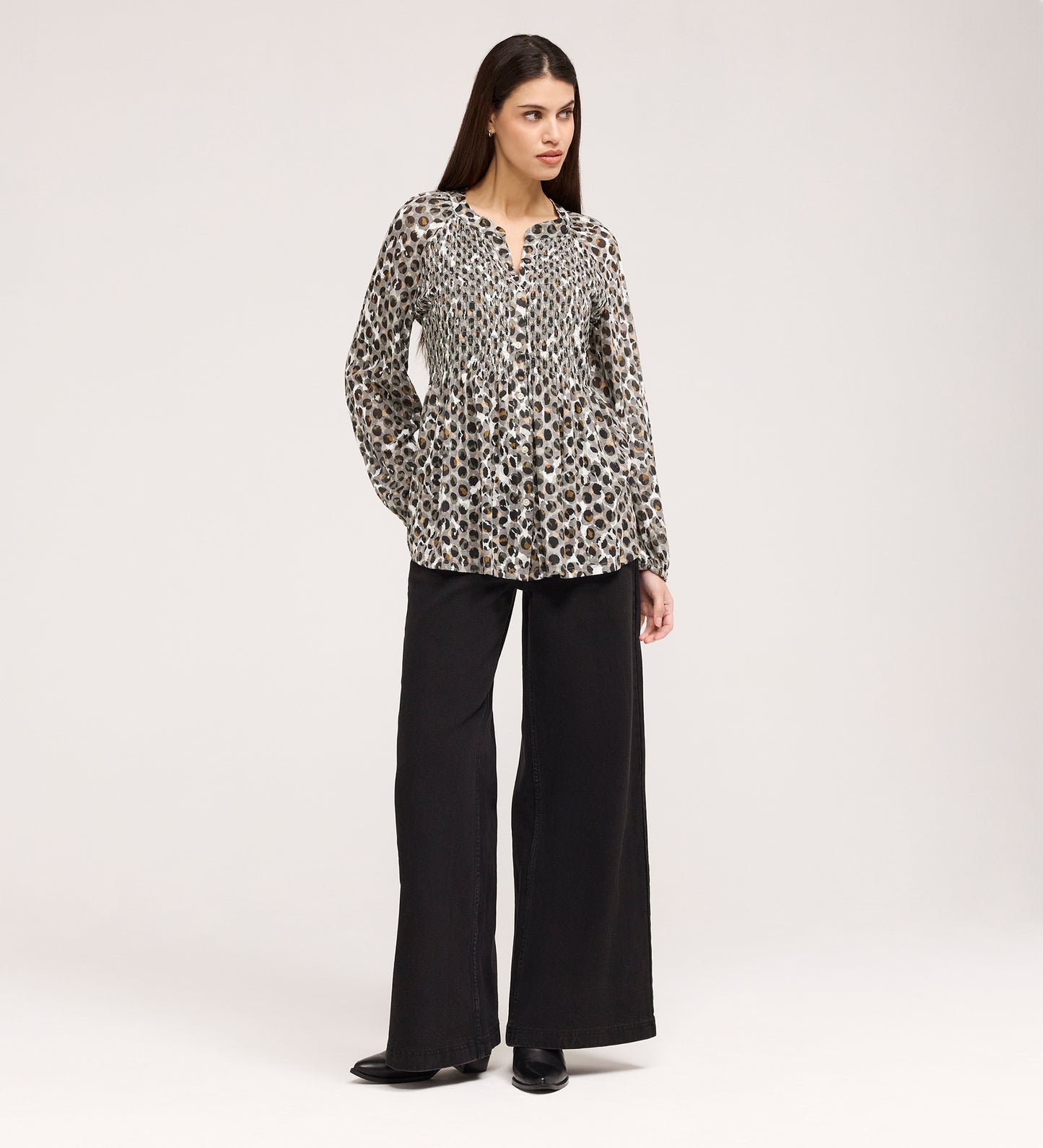 A woman with long, straight brown hair stands against a plain background, hands in pockets, wearing the Martha Khaki Animal Jersey Blouse—a loose, long-sleeve top with leopard print—paired with wide-leg black pants and black shoes. Subtle shirring adds texture.