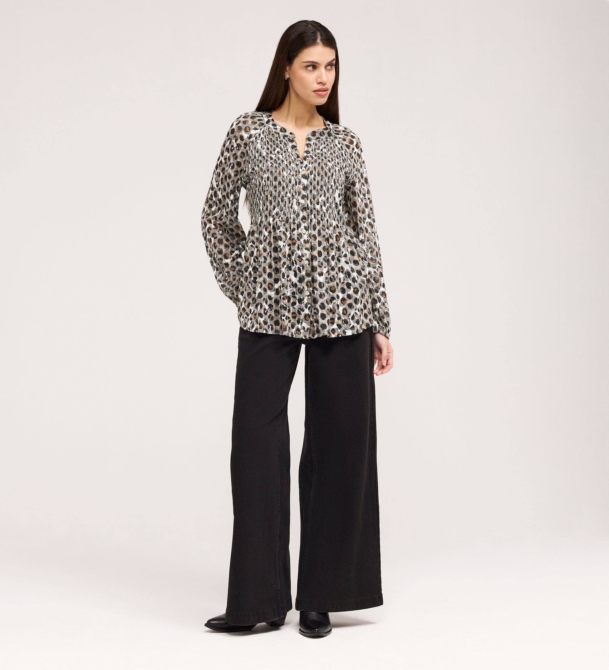 A woman with long, straight brown hair stands against a plain background, hands in pockets, wearing the Martha Khaki Animal Jersey Blouse—a loose, long-sleeve top with leopard print—paired with wide-leg black pants and black shoes. Subtle shirring adds texture.