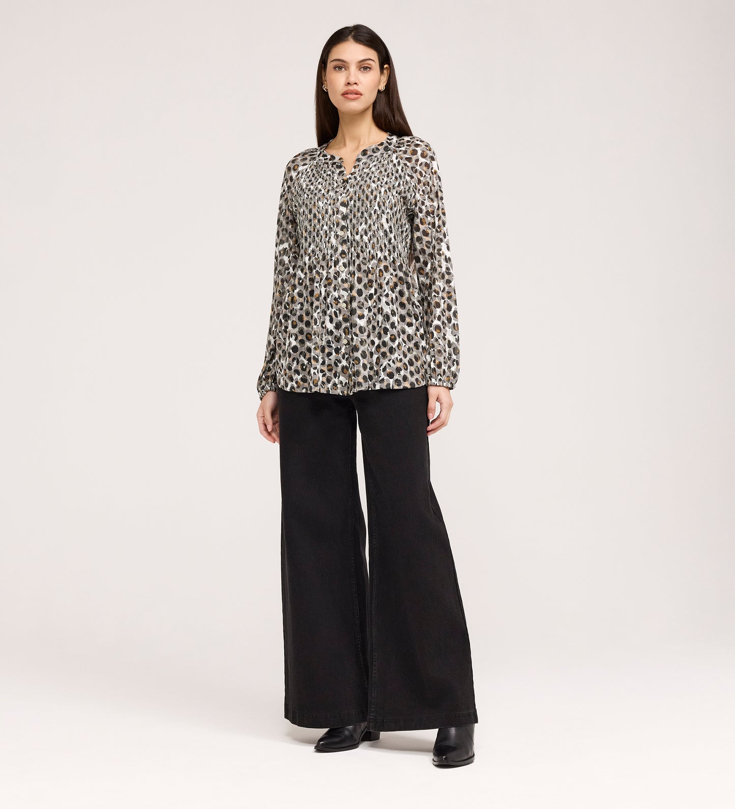 A woman stands facing forward against a plain light background, wearing the Martha Khaki Animal Jersey Blouse with black and beige spots, textured shirring, wide-leg black pants, and black shoes.