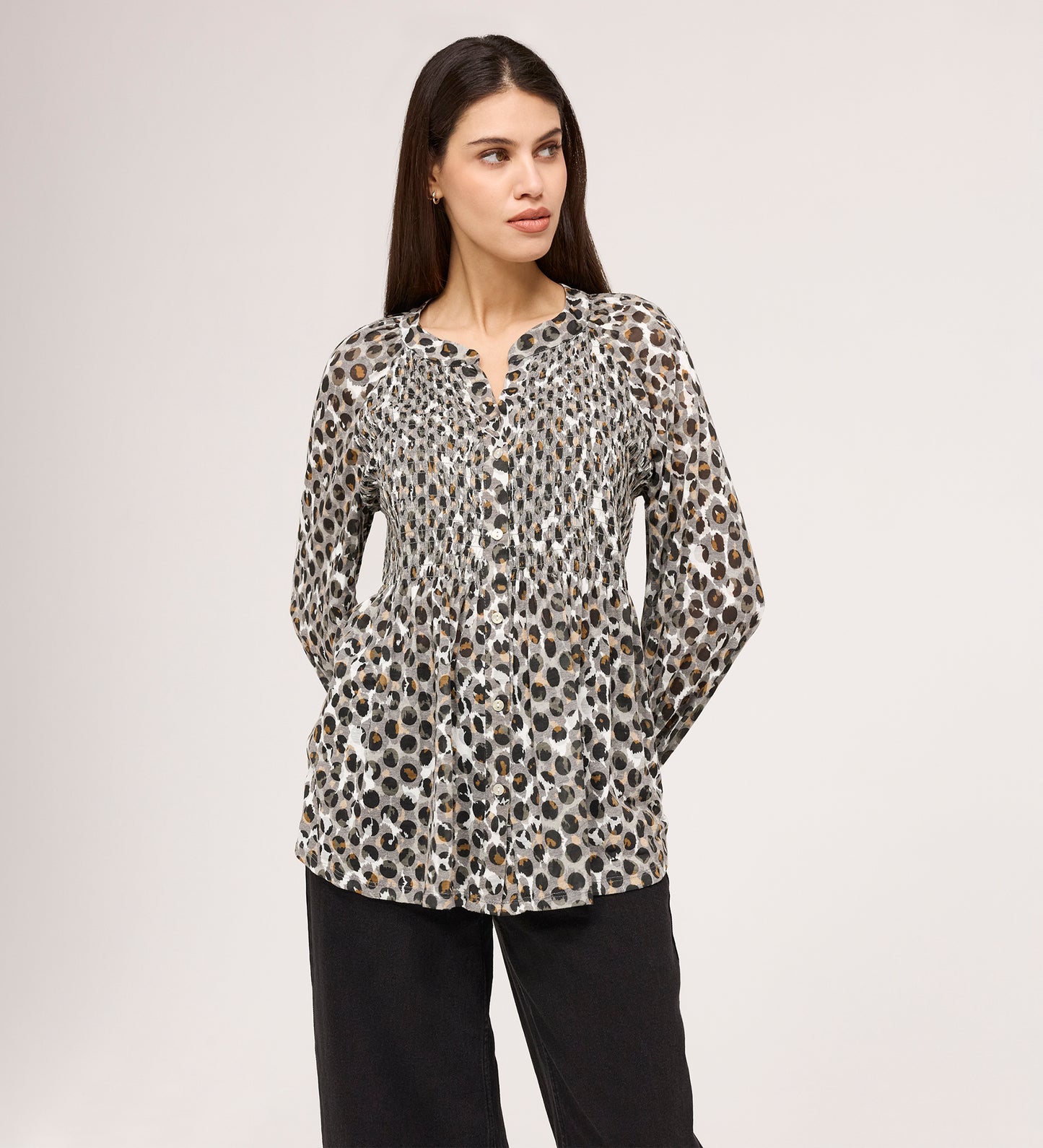 A woman with long brown hair looks to the side, wearing the Martha Khaki Animal Jersey Blouse—a long-sleeve, button-up top with a black, brown, and white animal print—paired with loose black pants against a plain background.