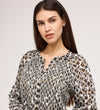 Woman with long dark hair wears the Martha Khaki Animal Jersey Blouse, featuring black, white, and brown spots with textured shirring, standing against a light gray background and looking at the camera neutrally.