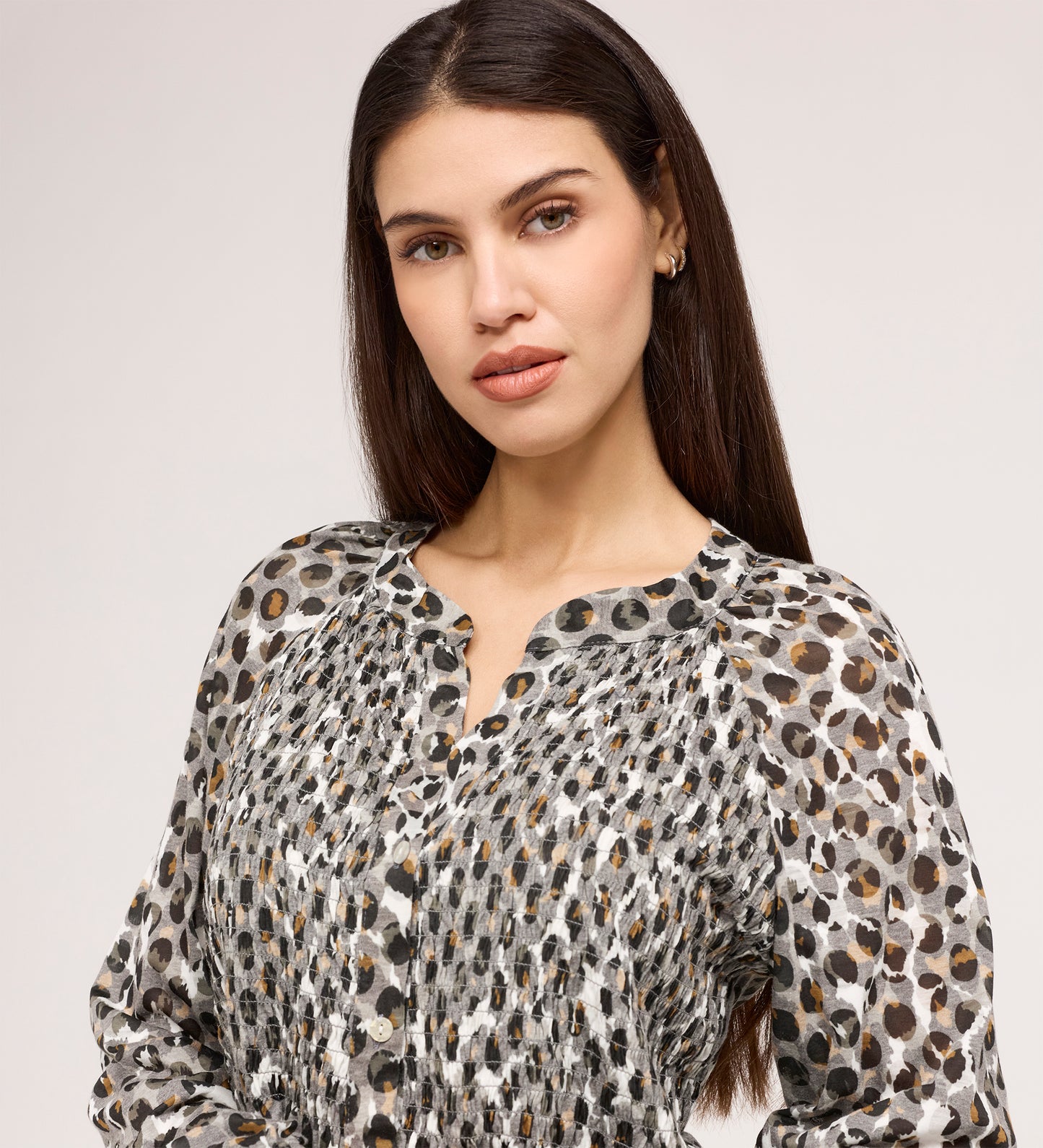 Woman with long dark hair wears the Martha Khaki Animal Jersey Blouse, featuring black, white, and brown spots with textured shirring, standing against a light gray background and looking at the camera neutrally.