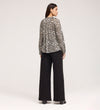 A woman faces away, wearing the Martha Khaki Animal Jersey Blouse with long sleeves and leopard print, paired with wide-leg black pants and platform shoes, set against a plain light background.