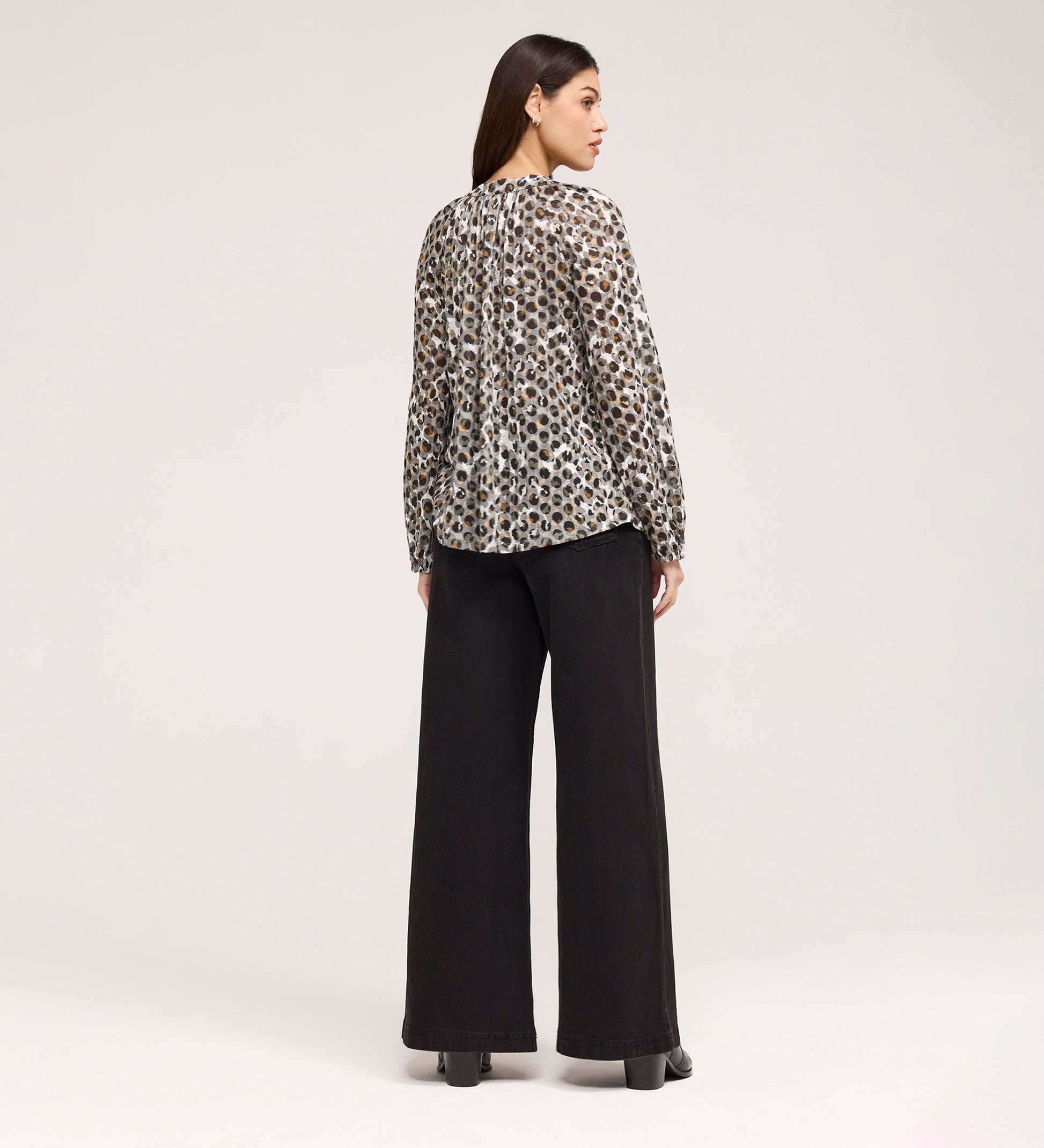 A woman faces away, wearing the Martha Khaki Animal Jersey Blouse with long sleeves and leopard print, paired with wide-leg black pants and platform shoes, set against a plain light background.