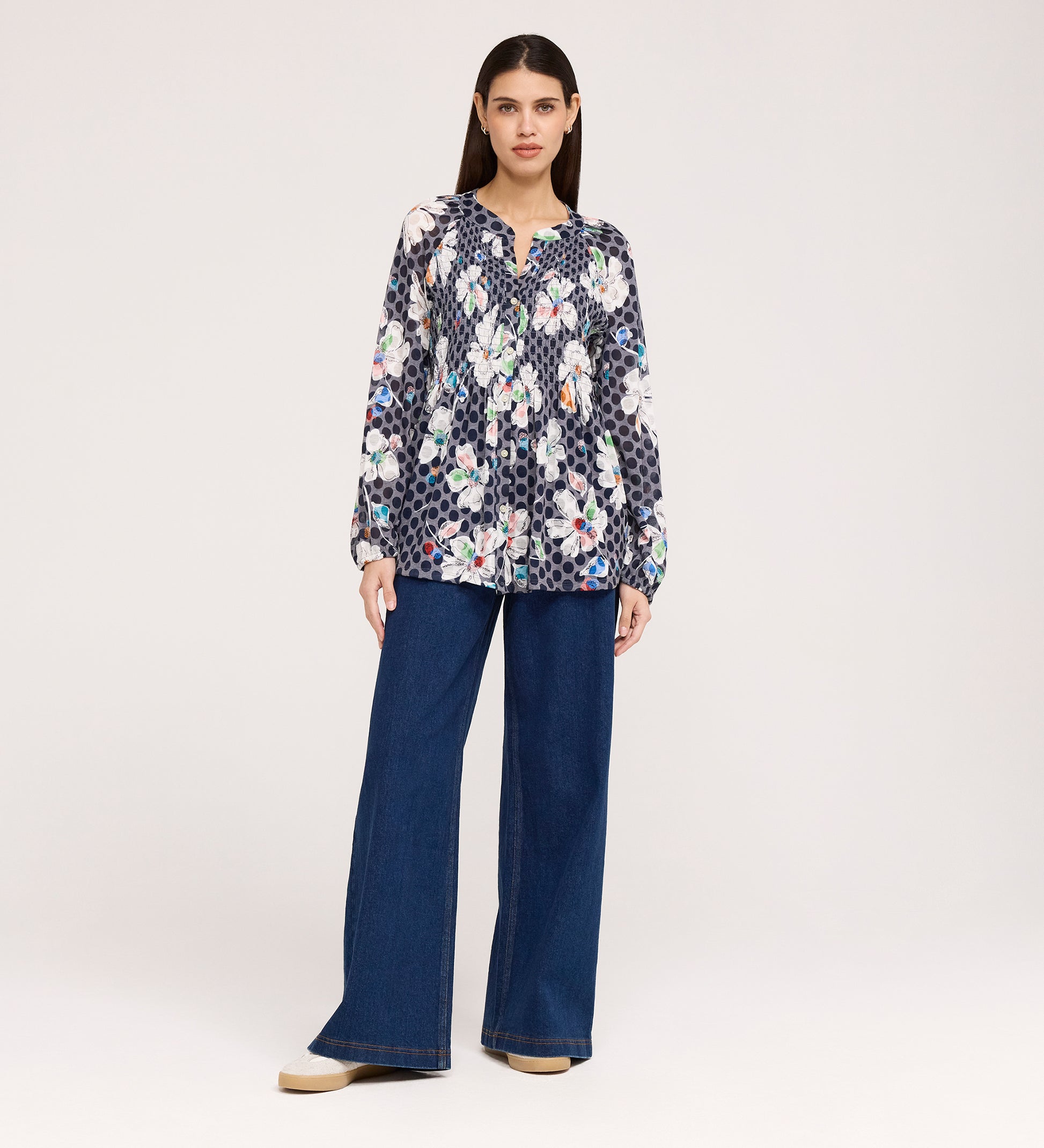 A woman stands against a plain background wearing the Martha Navy Flower Jersey Blouse, a long-sleeve top with floral and polka dot patterns and textured shirring, paired with wide-leg blue jeans and beige platform shoes.
