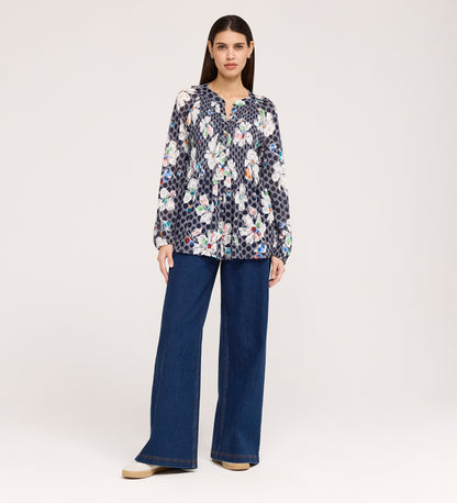 A woman stands against a plain background wearing the Martha Navy Flower Jersey Blouse, a long-sleeve top with floral and polka dot patterns and textured shirring, paired with wide-leg blue jeans and beige platform shoes.