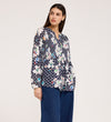 A woman in a Martha Navy Flower Jersey Blouse with textured shirring, floral and polka dot patterns, long sleeves, and blue wide-leg jeans stands against a white background, looking slightly left with a neutral expression.
